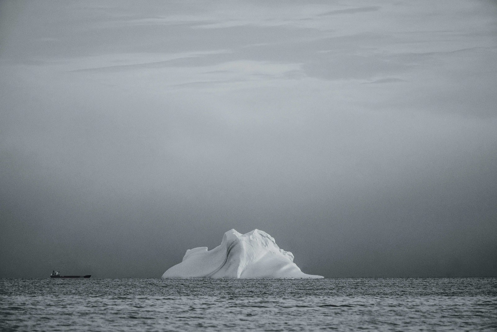 An iceberg and a distant cargo vessel under a gray sky